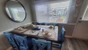 A dining area with a wooden table set for four, blue chairs, a mirror on the wall, and a window with blinds. - Deep-Water Canal Front Coastal Cottage Only Steps to the Beach & Pet Friendly!  (Surf City)