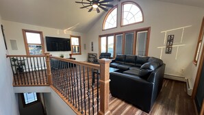 A spacious living area with a black leather sofa, wooden flooring, a wooden staircase with a railing, and large windows allowing natural light. - Newly Renovated 4BR 2BA Ocean Block Bay View Ship Bottom Home Central Location  (Ship Bottom)