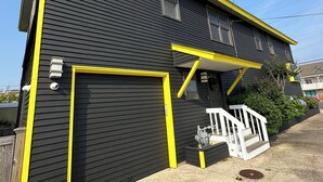 A two-story building with a black exterior and yellow trim, featuring a garage door and a small porch with steps. - Newly Renovated 4BR 2BA Ocean Block Bay View Ship Bottom Home Central Location  (Ship Bottom)