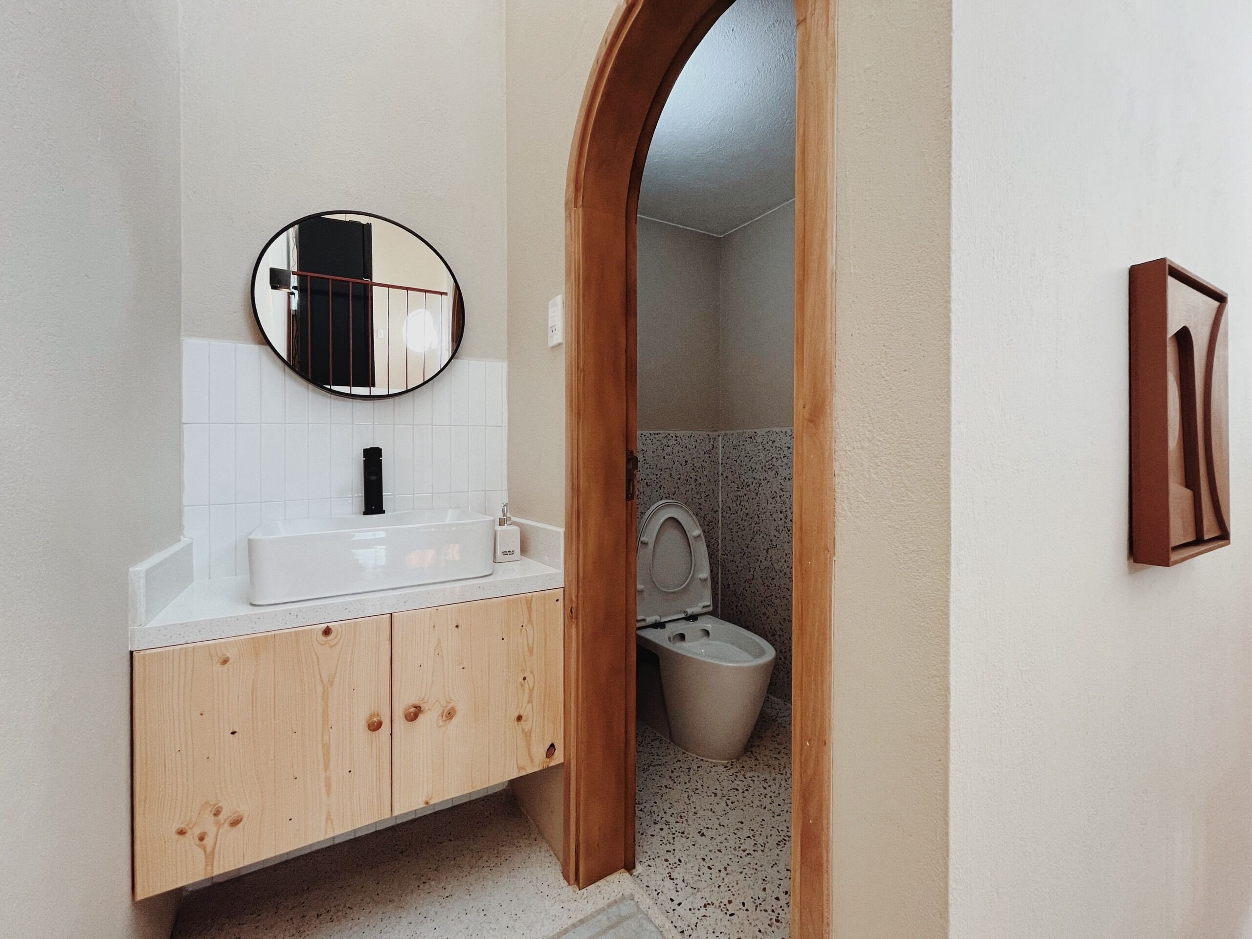 Une salle de bain avec un lavabo blanc, un meuble-lavabo en bois, un miroir rond et des toilettes.