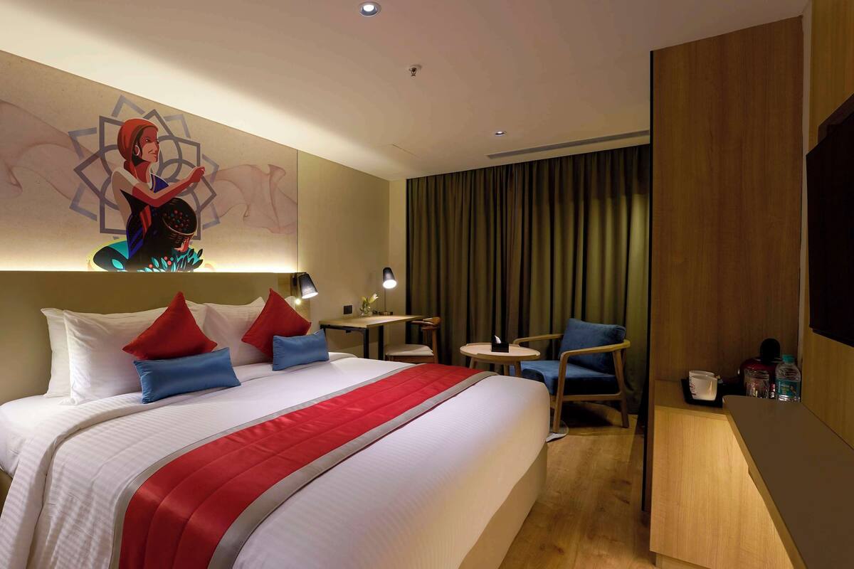 Une chambre d’hôtel avec un grand lit, deux tables de chevet, un bureau avec une chaise, une télévision et un mur orné d’un motif géométrique.