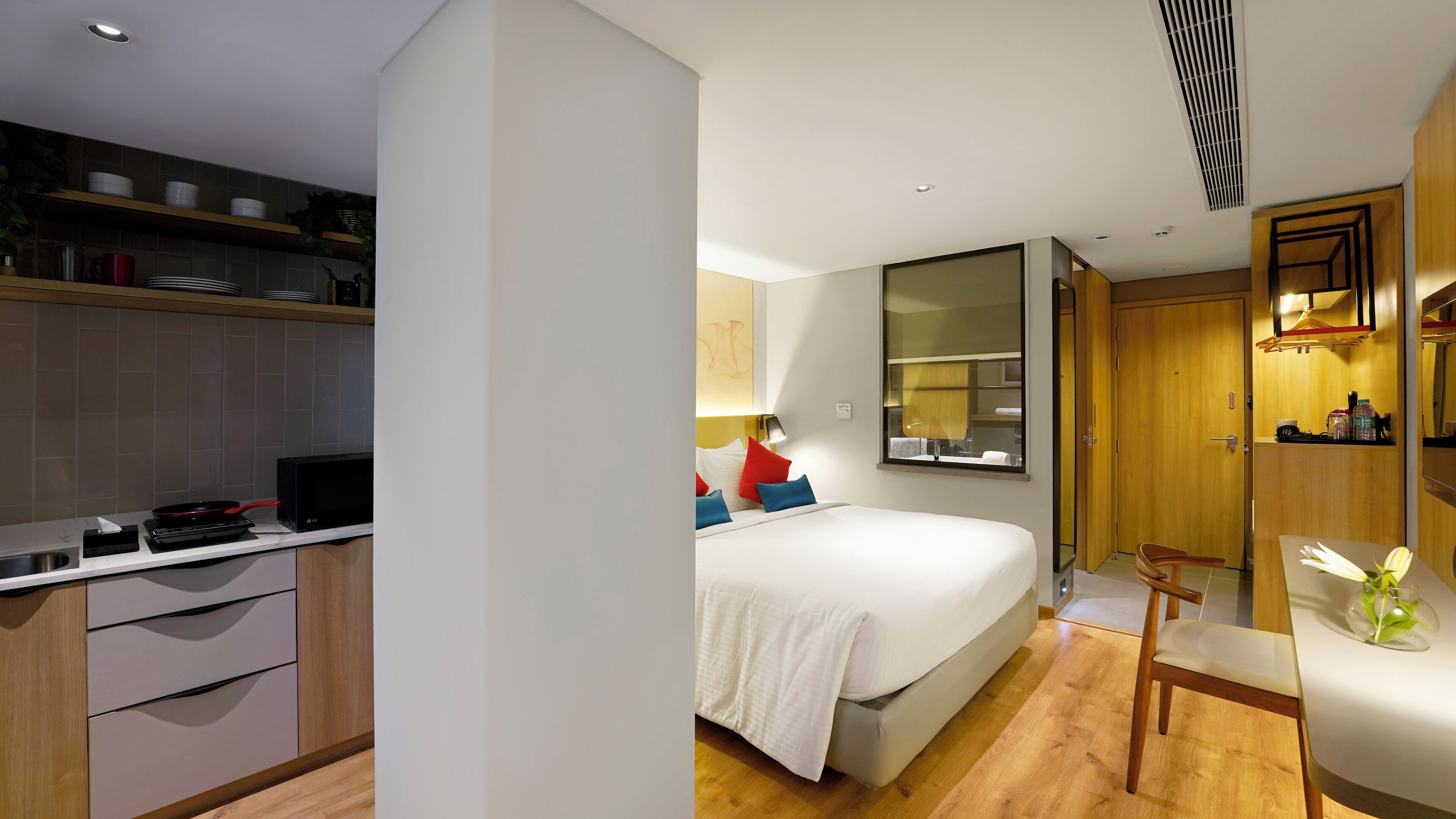 A modern hotel room with a kitchenette, a bed with white bedding, a bedside table with a lamp, and a wooden floor.