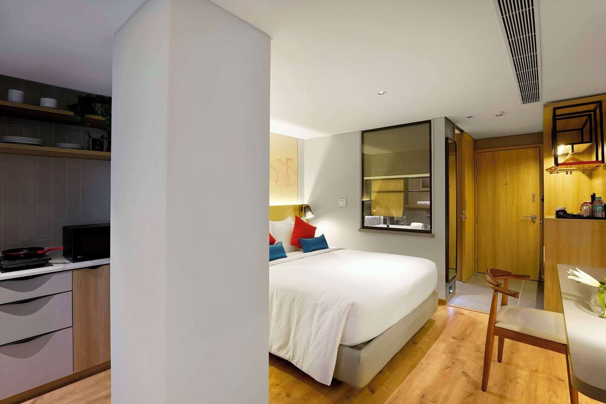 Une chambre d’hôtel moderne dotée d’une kitchenette, d’un lit agrémenté d’oreillers colorés, d’une table de chevet avec une lampe et d’un grand miroir.