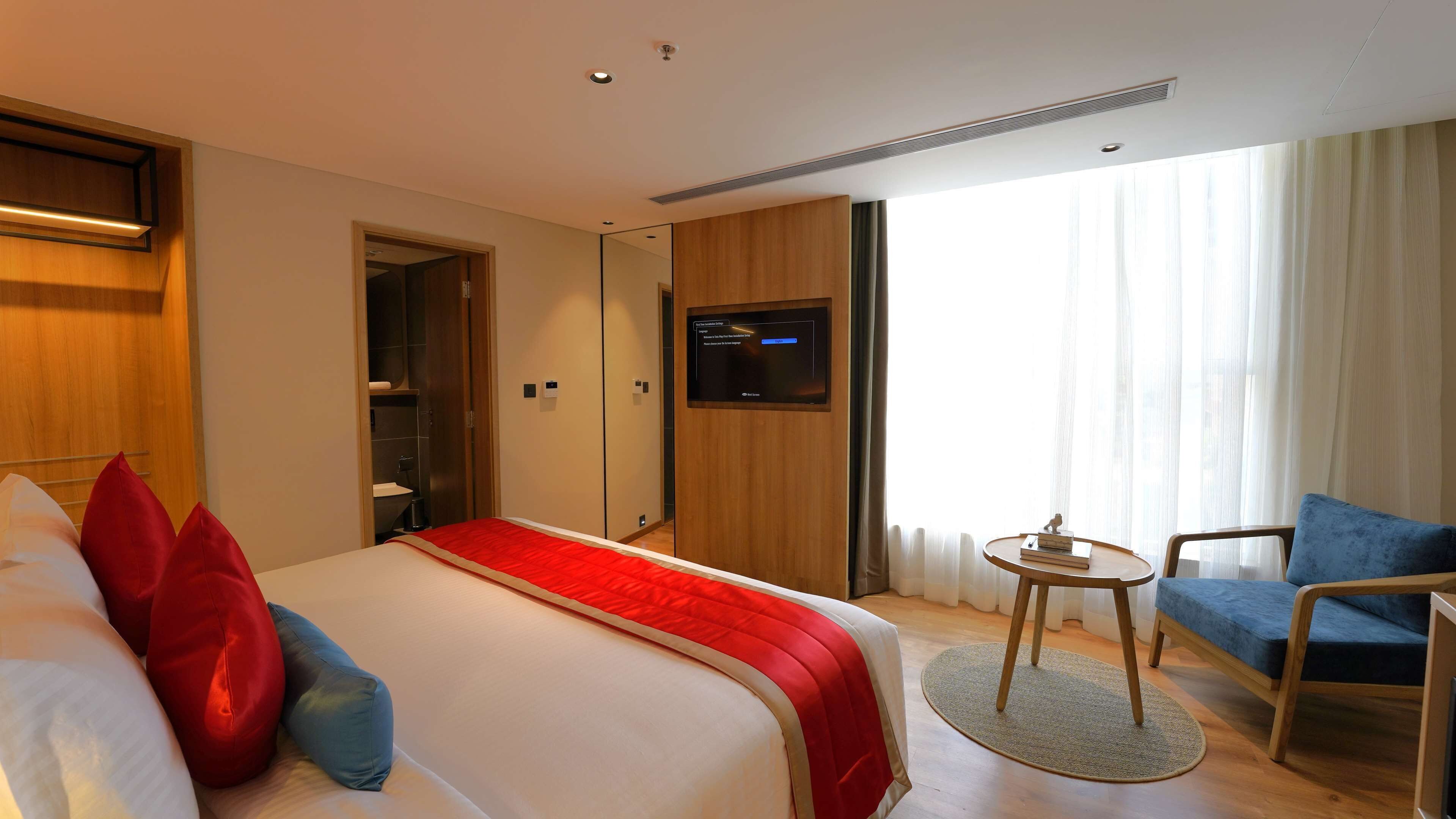 Habitación de hotel con cama, televisor montado en la pared, escritorio con silla y ventana con cortinas.