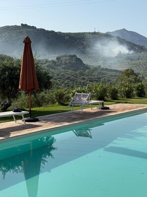 Ein Swimmingpool mit Liegestühlen und Sonnenschirm, umgeben von Gr ünpflanzen und Bergen im Hintergrund.