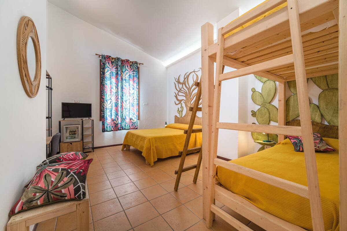 A room with two beds, a wooden ladder, a desk with a chair, a television, and a window with colorful curtains.
