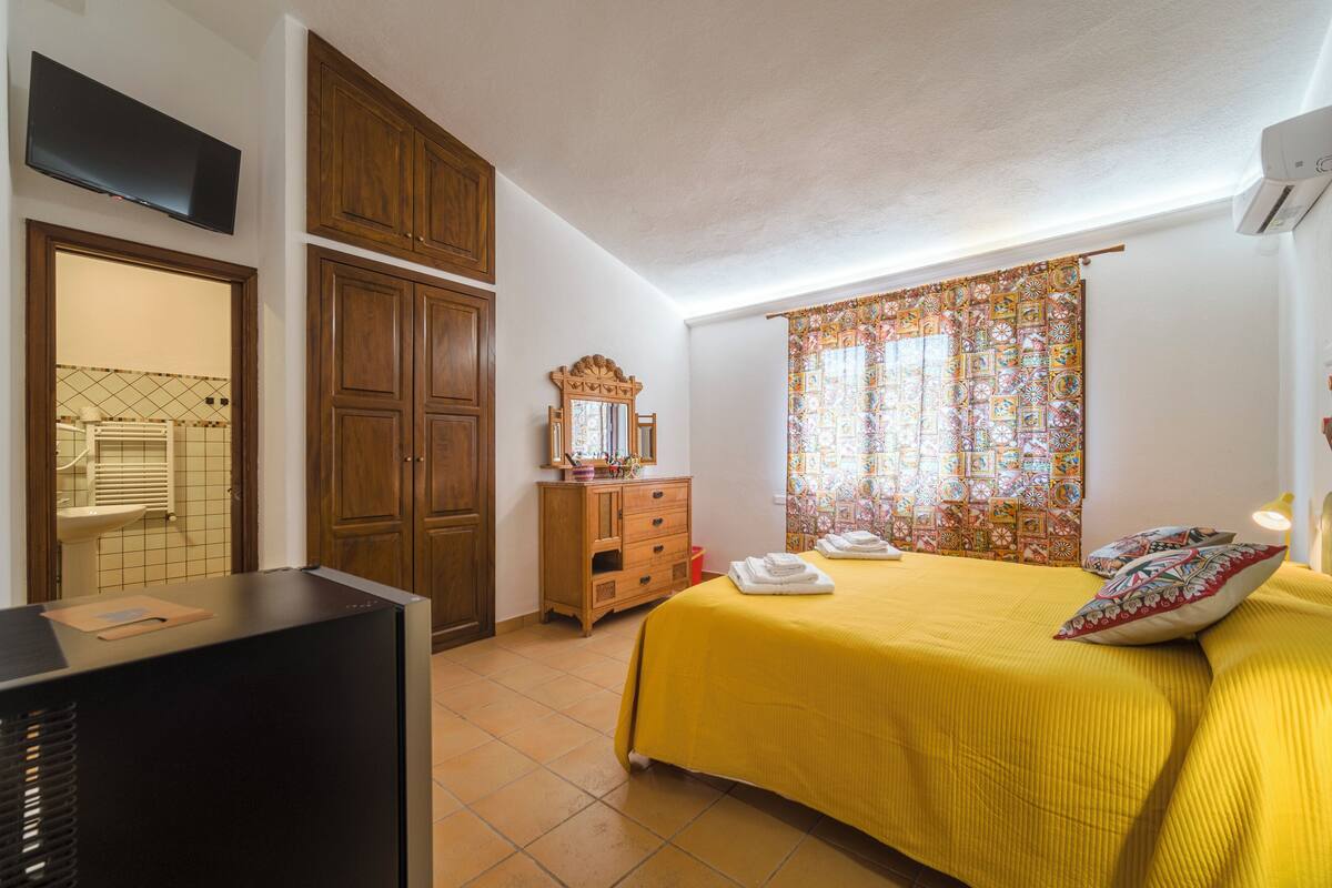 A bedroom with a bed, a dresser, a television, and a window with patterned curtains.