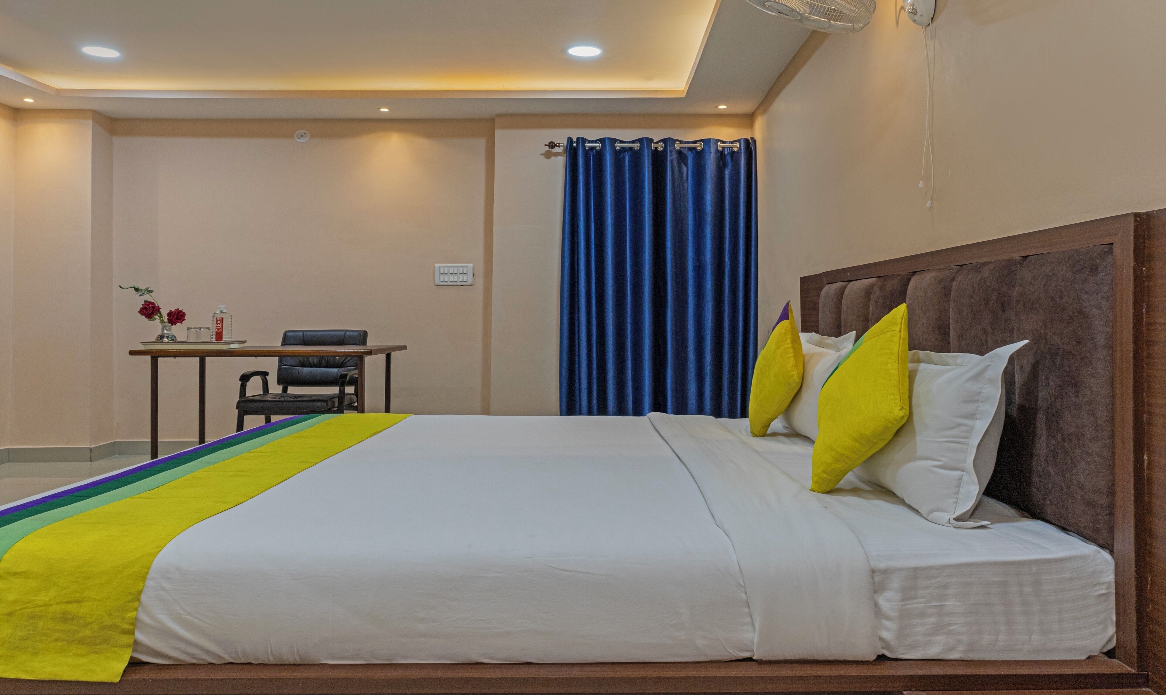 A hotel room with a large bed, a desk with a chair, a blue curtain, and a ceiling fan.