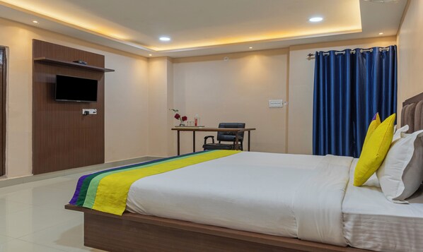 Habitación de hotel con cama, escritorio, silla, televisión y ventana con cortinas.