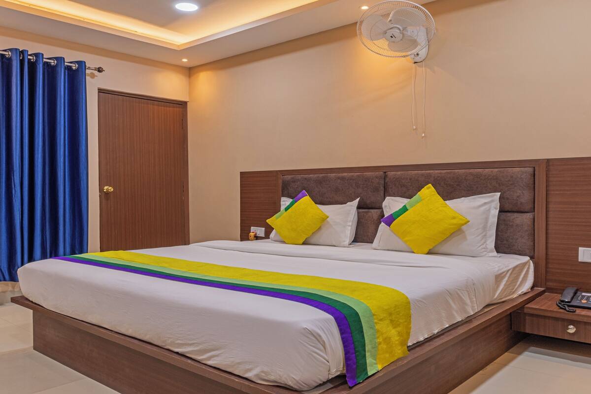 A hotel room with a bed, a bedside table with a phone, a wall-mounted fan, and a door with a curtain.