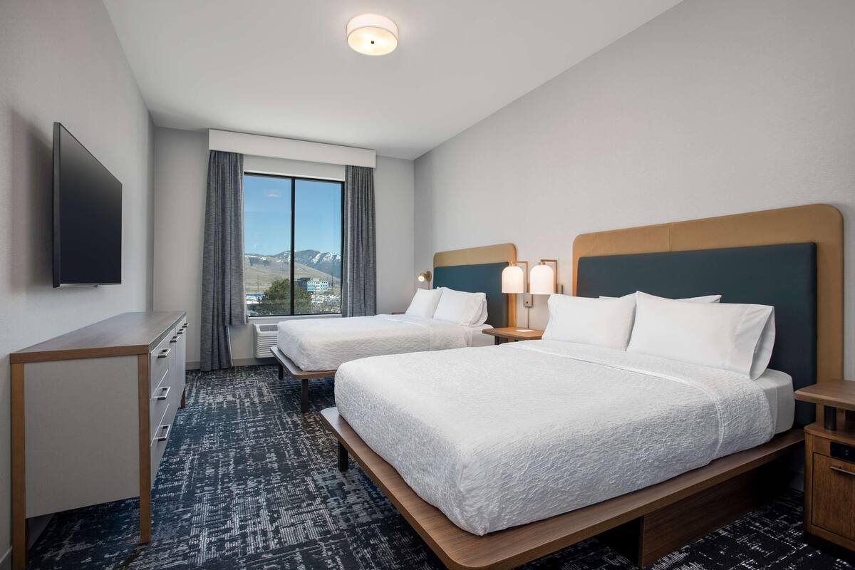 Habitación de hotel con dos camas, un escritorio, un televisor y ventana con vista a la montaña.