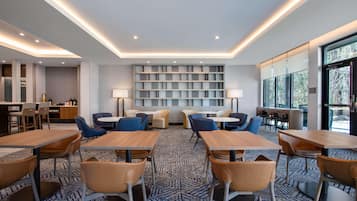 A modern dining area with tables and chairs, a bookshelf, and a view through large windows.