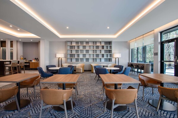 A modern dining area with tables and chairs, a bookshelf, and a view through large windows.