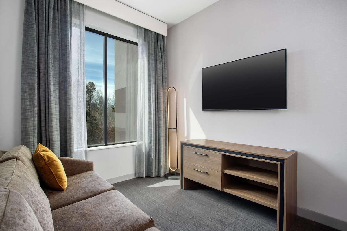 A modern hotel room with a large window, a flat-screen TV mounted on the wall, a wooden console table, and a sofa with a mustard-colored pillow.