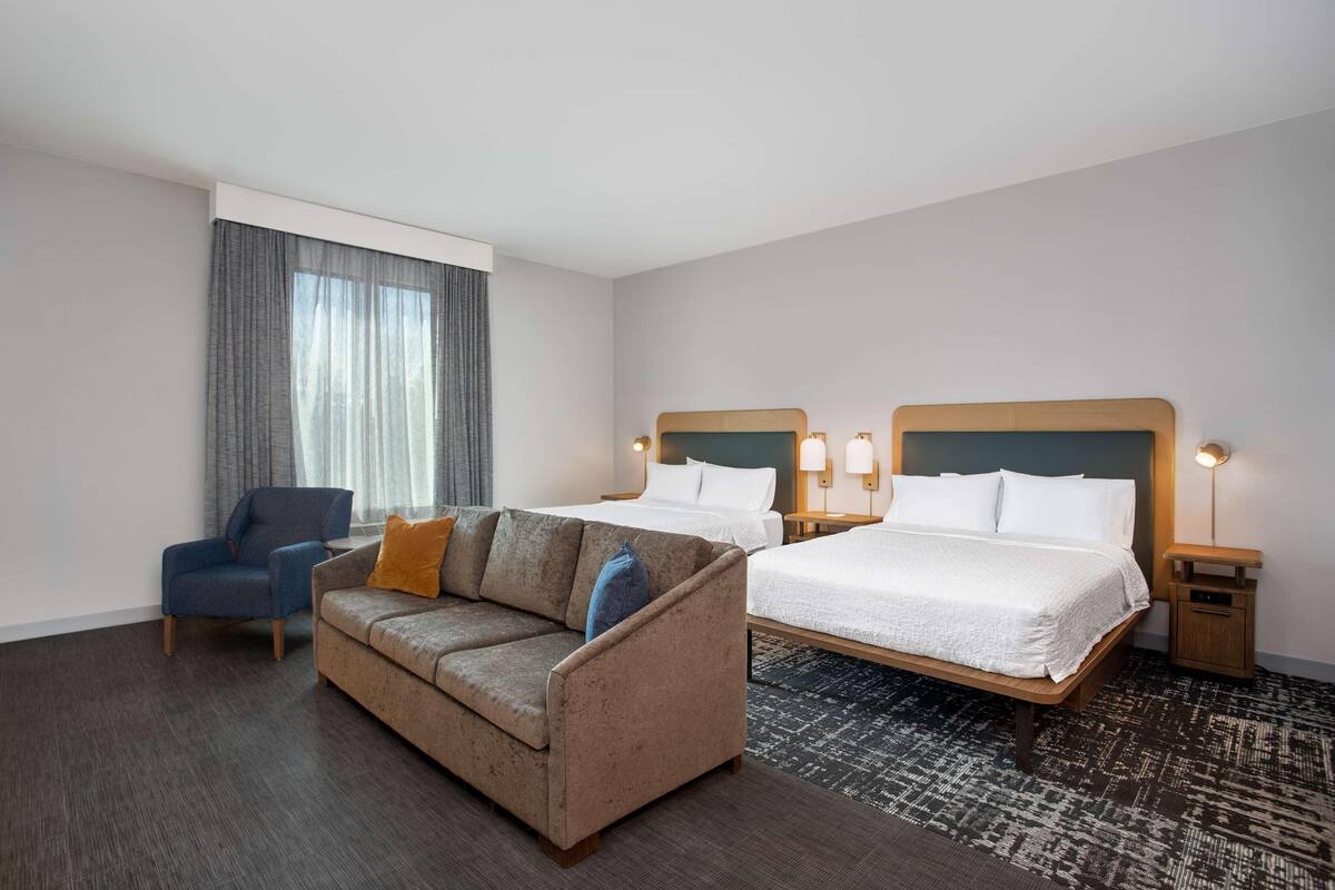 Una habitación de hotel con sofá, dos camas, una silla y una mesita de noche con lámparas.