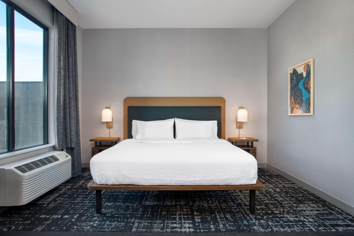 A hotel room with a large bed, two bedside lamps, a window with curtains, a wall-mounted air conditioner, and a painting on the wall.