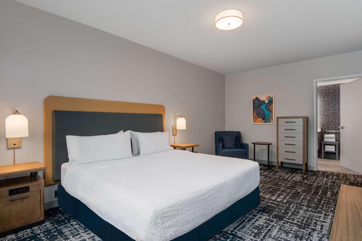 Habitación de hotel con una cama grande, mesitas de noche, una silla, un aparador y un cuadro en la pared.
