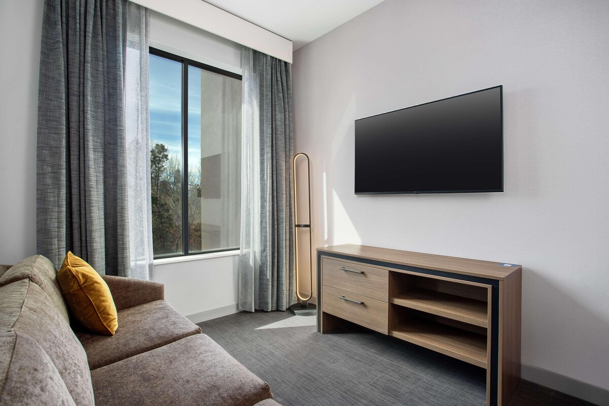A modern hotel room with a large window, a flat-screen TV mounted on the wall, a wooden console table, and a sofa with a mustard-colored pillow.