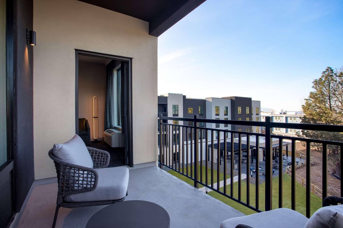 Un balcón con una silla de mimbre y una mesa, una puerta que da a una habitación interior, y vistas de edificios modernos y una zona ajardinada.