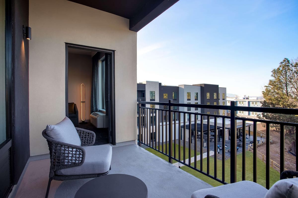 A balcony with a wicker chair and a table, a door leading to an interior room, and a view of modern buildings and a grassy area.