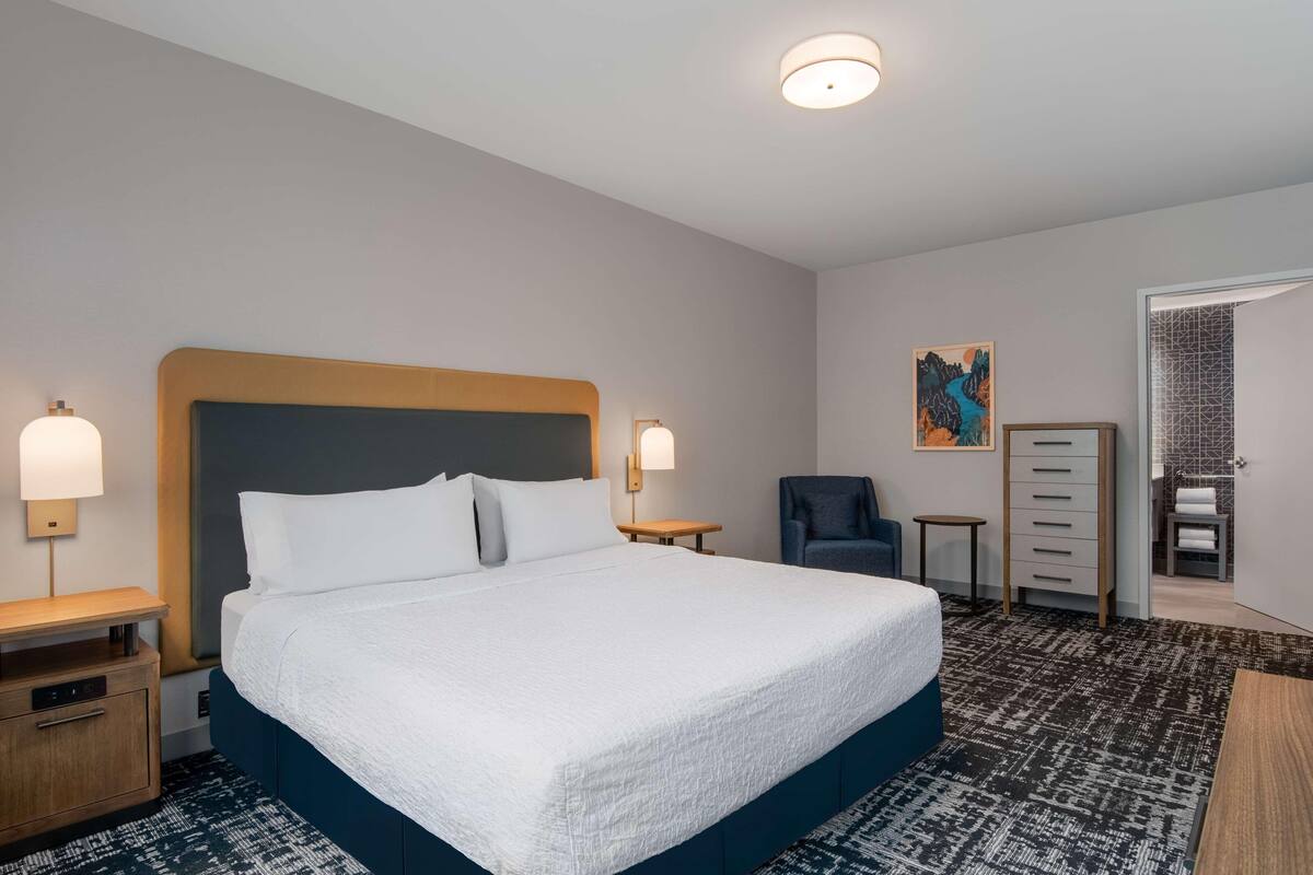 Habitación de hotel con una cama grande, mesitas de noche, una silla, un aparador y un cuadro en la pared.