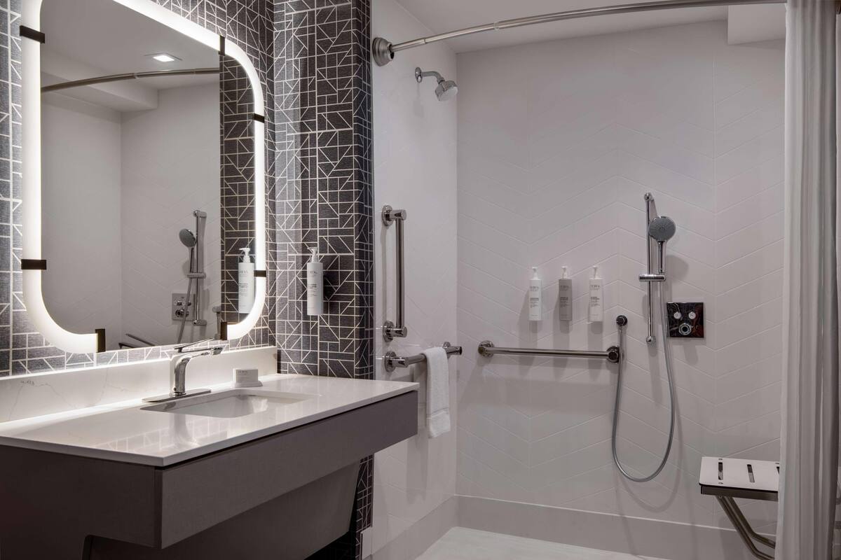 Un baño moderno con un lavamanos blanco, un espejo grande y una zona de ducha con cabezal de ducha manual.