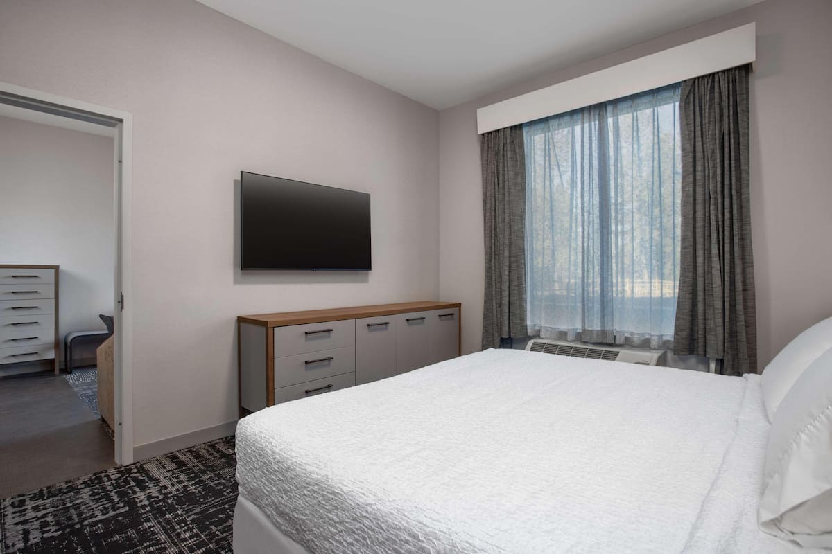 Habitación de hotel con cama, televisión montada en la pared, ventana con cortinas, cómoda y suelo alfombrado.