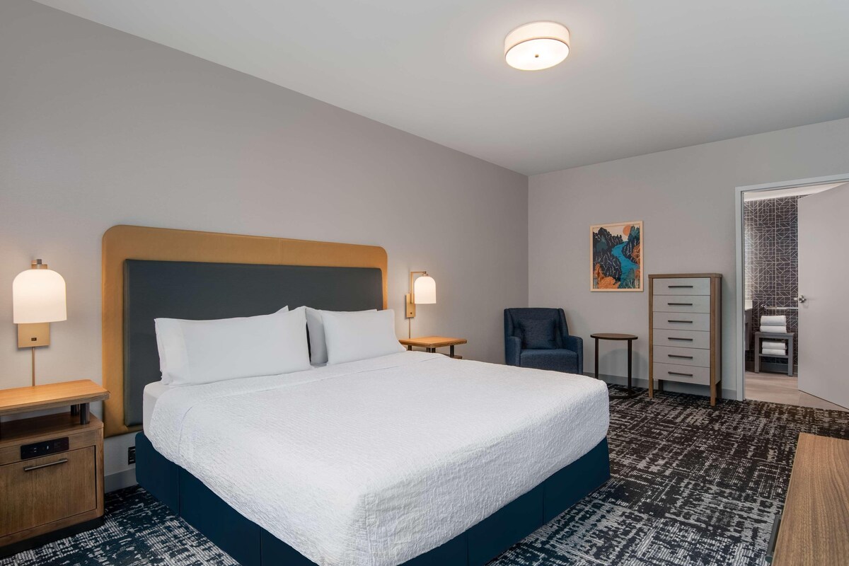 Ein Hotelzimmer mit einem großen Bett, Nachttischen, einem Sessel, einer Kommode und einem Bild an der Wand.
