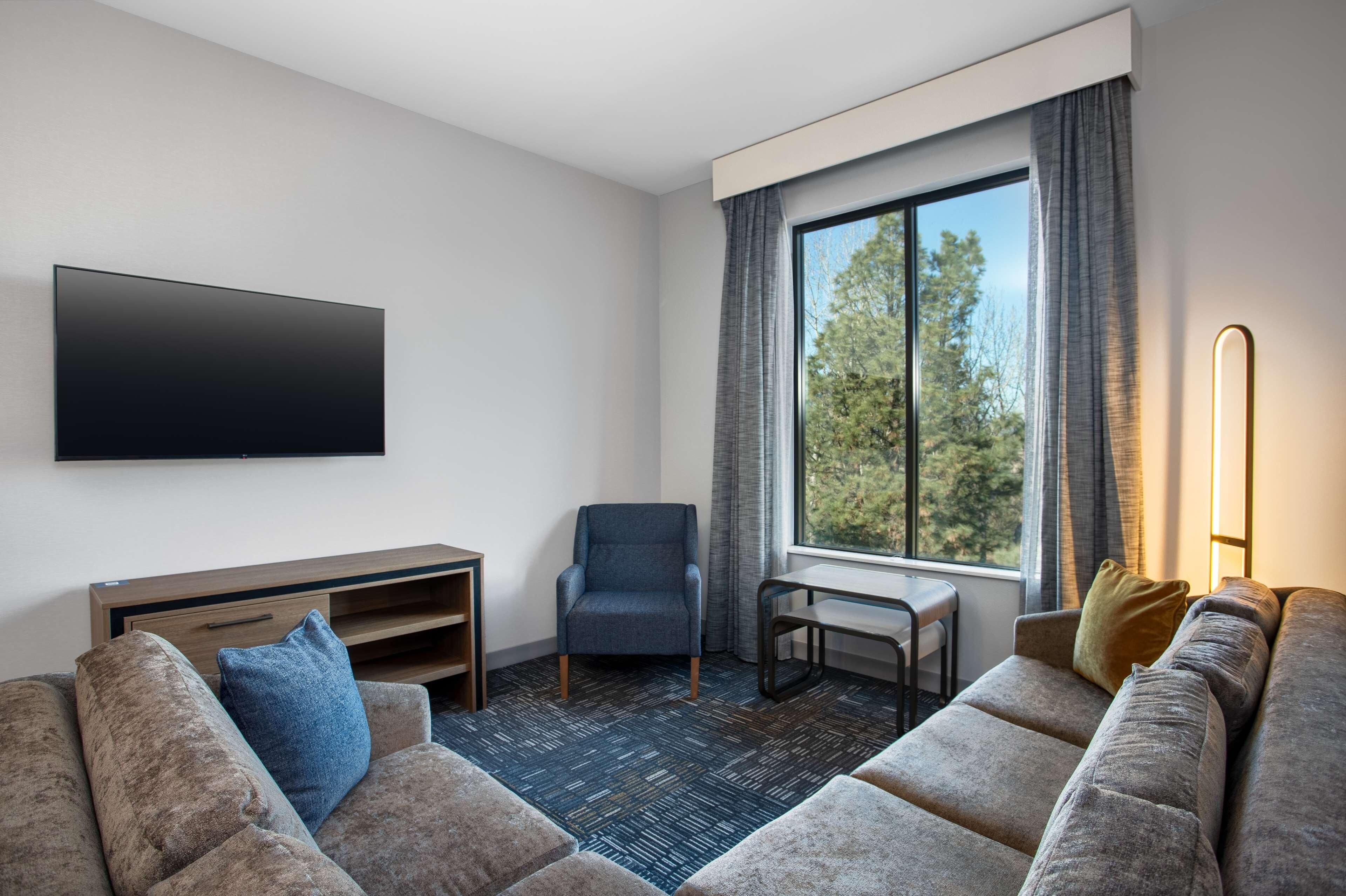 A modern hotel room with a large window, a flat-screen TV, a sofa, a chair, and a small table.