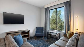 A modern hotel room with a large window, a flat-screen TV, a sofa, a chair, and a small table.