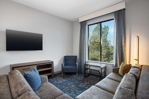 A modern hotel room with a large window, a flat-screen TV, a sofa, a chair, and a small table. - Homewood Suites by Hilton Missoula (Missoula)