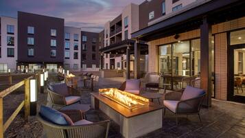 A modern outdoor seating area with a fire feature, wicker furniture, and a building with multiple floors and large windows.
