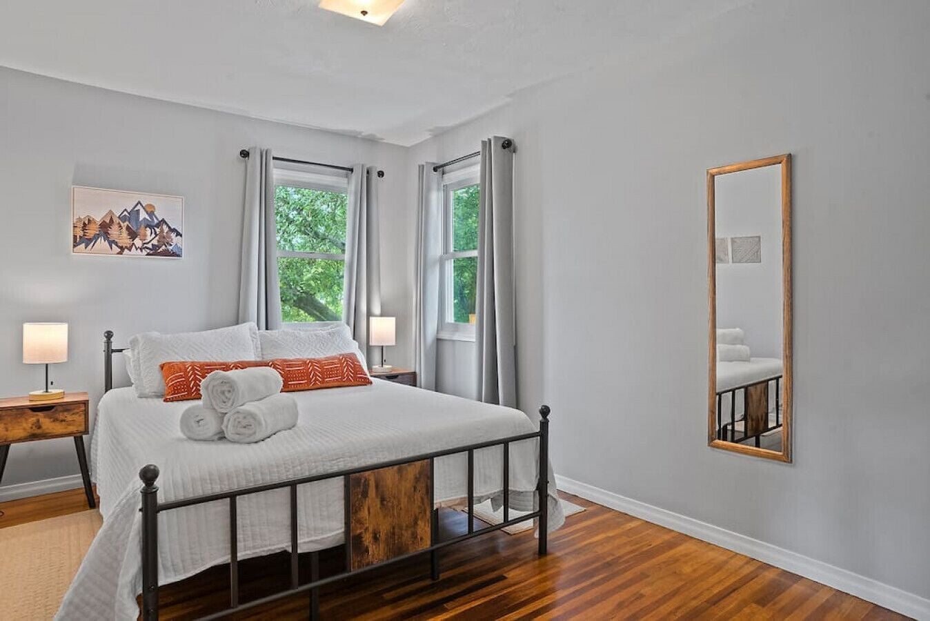 A neatly arranged bedroom with a bed, two bedside tables, a mirror, and a window with curtains.