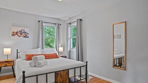 Un dormitorio ordenado con cama, dos mesitas de noche, un espejo y una ventana con cortinas.