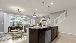 A modern kitchen with a central island, pendant lighting, and a view to a living area with a sofa and coffee table.