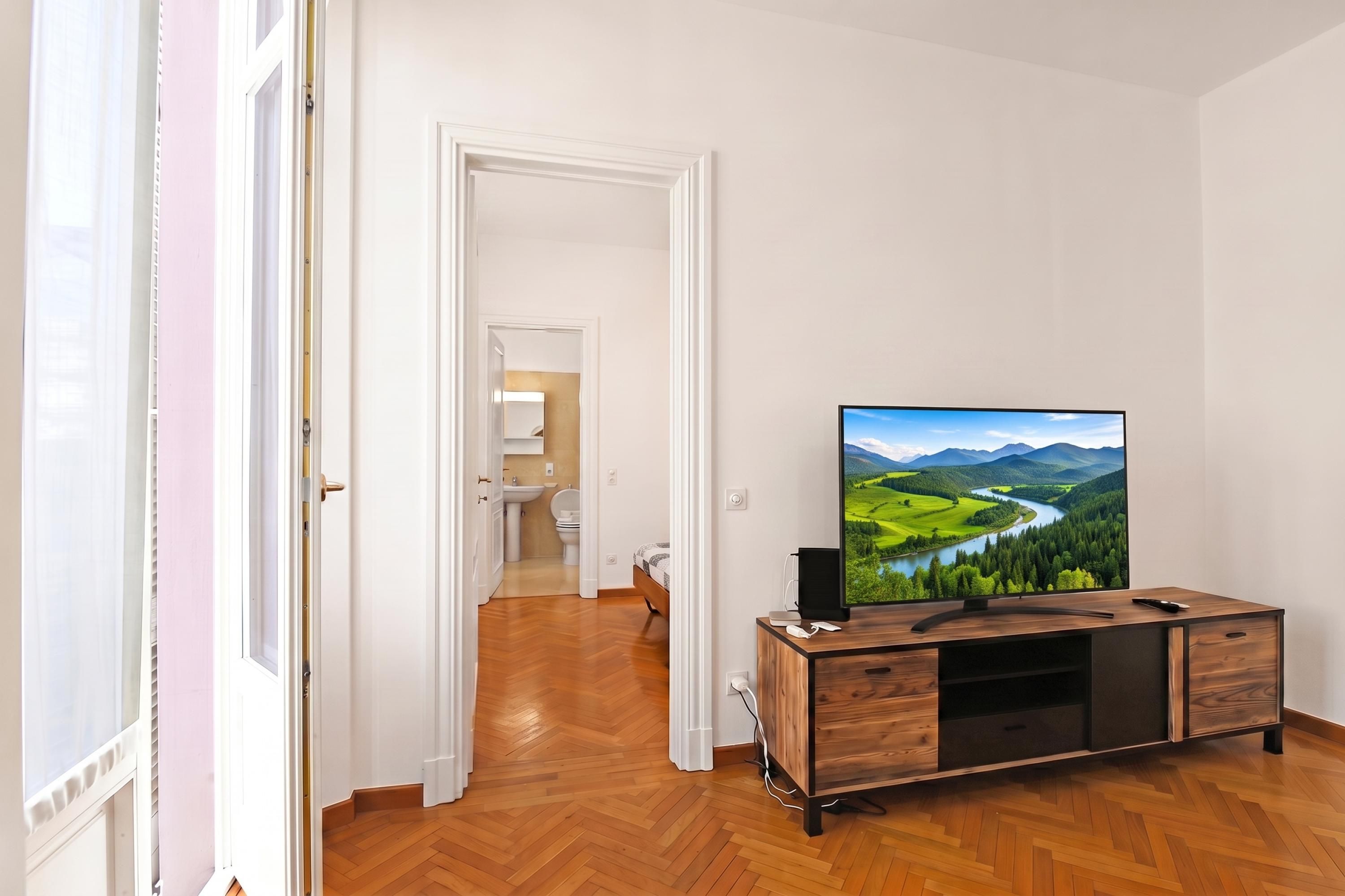 A hotel room with a wooden floor, a TV on a wooden stand, and a view of a landscape through the window.