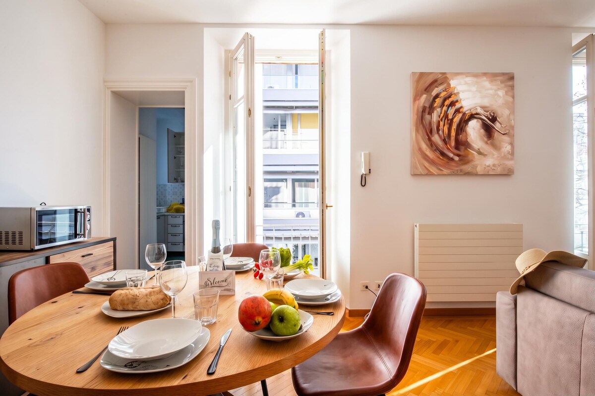 A dining area with a round table, chairs, a microwave, a painting, and a view of the outside.