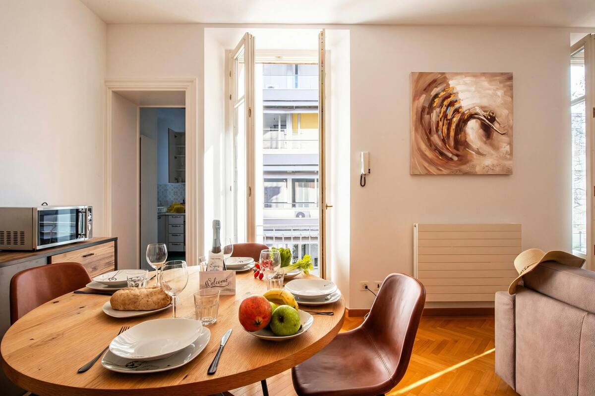 Un comedor con una mesa redonda preparada para cuatro personas, un sofá, un cuadro en la pared y vistas del edificio exterior.
