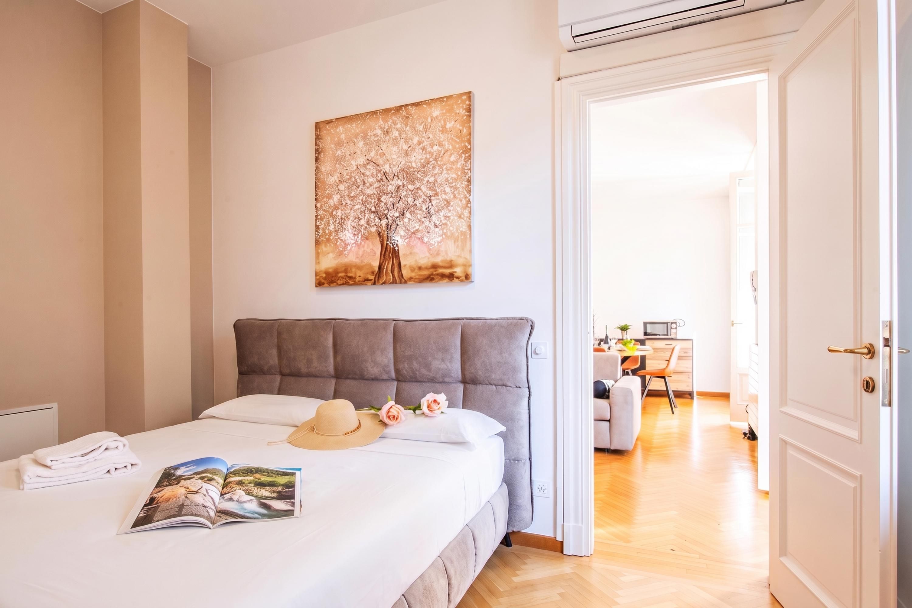 A bedroom with a bed, a bedside table, a magazine, a hat, and a painting on the wall.