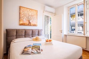 A hotel room with a large bed, a painting of a tree, a window with a view of buildings, and a magazine on the bed.