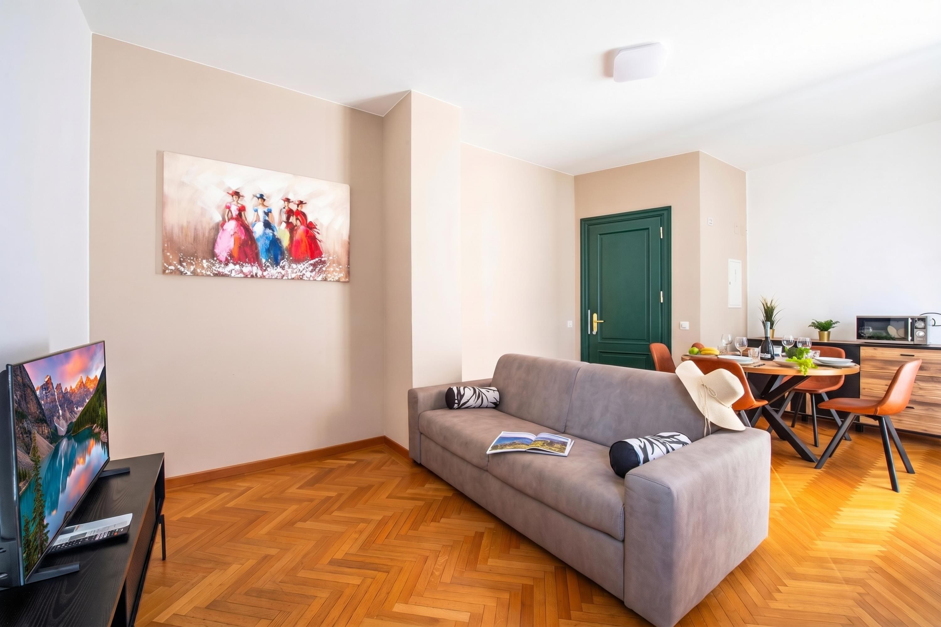 Uma sala de estar moderna com um sofá cinza, um quadro na parede, uma área de jantar com uma porta verde e uma TV em um suporte de madeira.