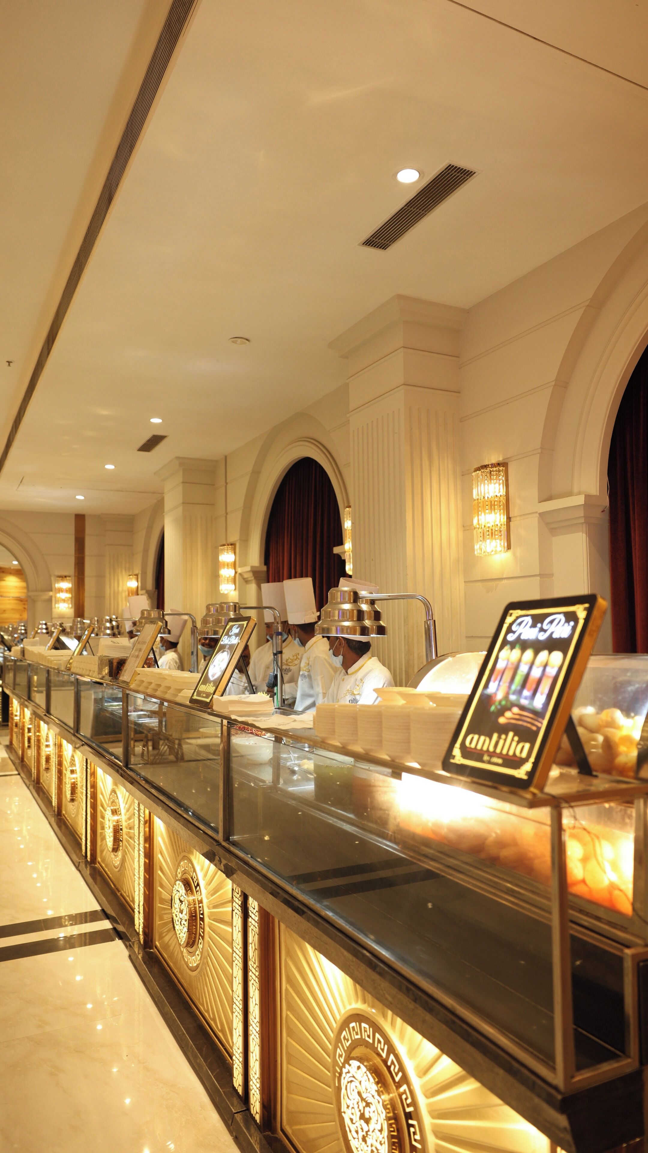 A luxurious hotel lobby with a long counter displaying various food items, elegant chandeliers, and a grand arched doorway.