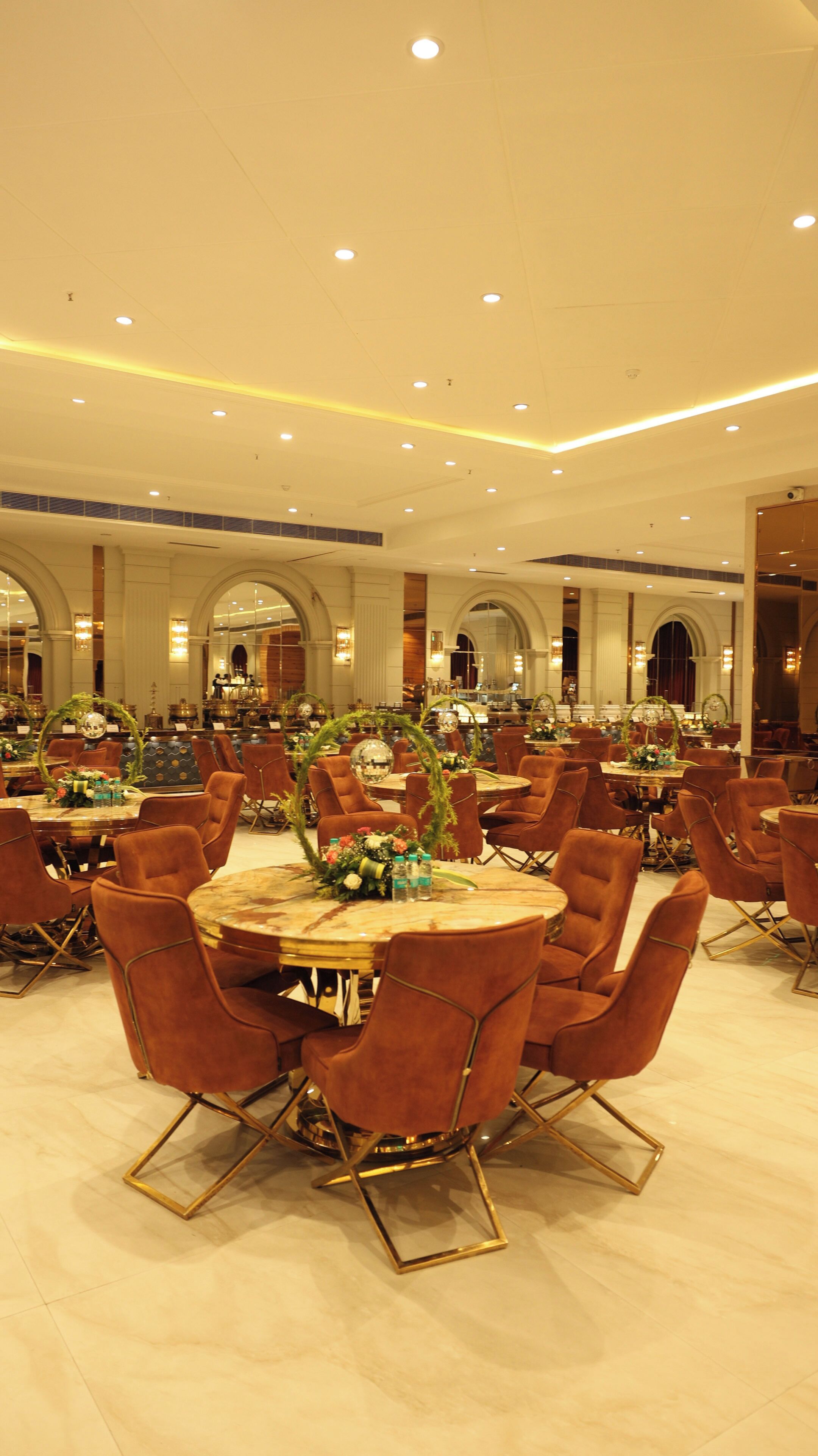 A spacious banquet hall with round tables, brown upholstered chairs, and a high ceiling with recessed lighting.