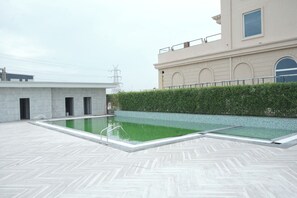 A rooftop swimming pool with a herringbone tile floor, surrounded by a low hedge and a building with arched windows. - Antilia By Zion (Sonepat)