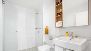 Et stilfuldt badeværelse med en glasvægsbrusekabine, et hvidt toilet, en vask med et spejl over, og en hylde med toiletartikler.