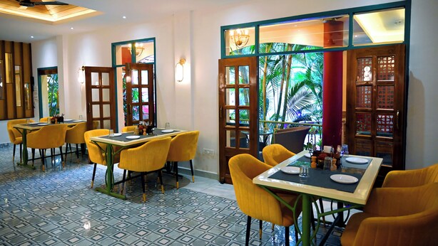 A dining area with yellow chairs, a table set for two, and a view of tropical plants through a glass door. -  Woods Inn Resort (Airport Hotel) (Bhopal)