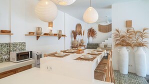 A modern kitchen with white countertops, a built-in microwave, and a dining area with a long table set for four. There are hanging paper lanterns, decorative vases, and a staircase leading to an upper level. - Villa Madissa by Villa Finder (Canggu)