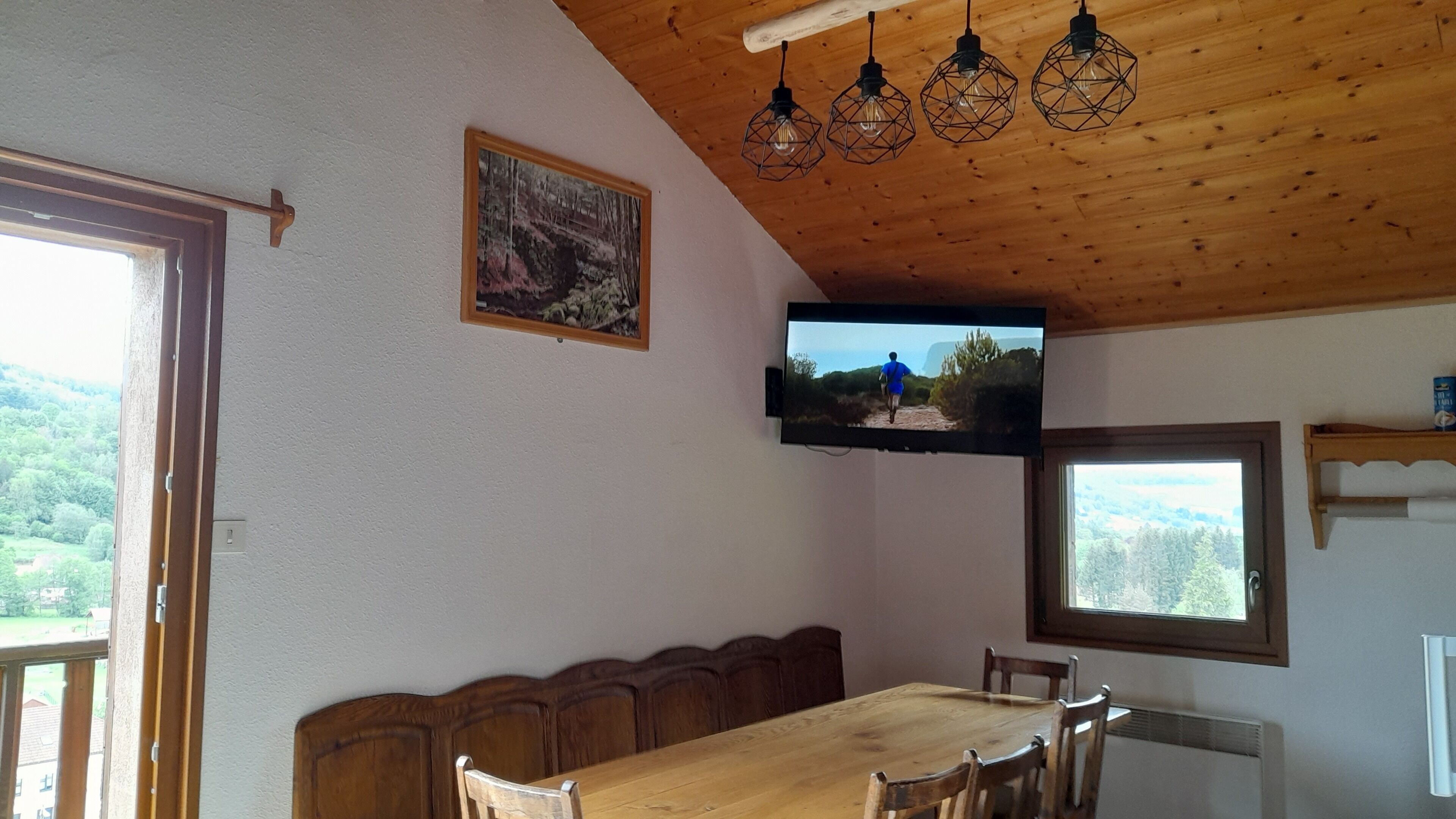 A dining area with a wooden table and chairs, a television mounted on the wall, and a window with a view of greenery.