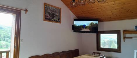 A dining area with a wooden table and chairs, a television mounted on the wall, and a window with a view of greenery.