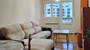 Una sala de estar con un sofá, una mesita y una silla. Se ven edificios a través de las ventanas.